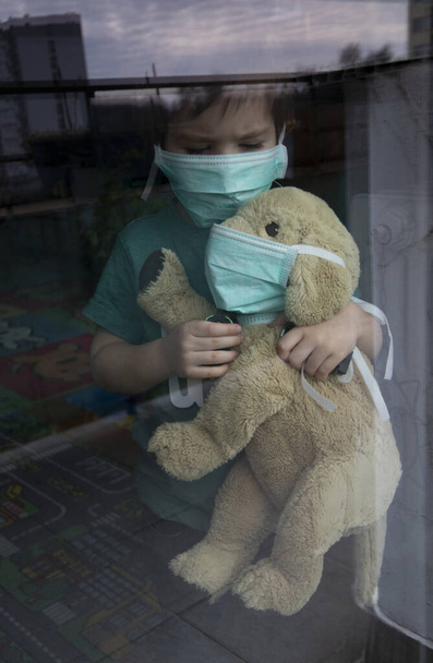 Coronavirus, Covid-19, Boy a jeho plyšové štěně v ochranných maskách se dívají oknem, doma v karanténě  - Fotografie, Obrázek