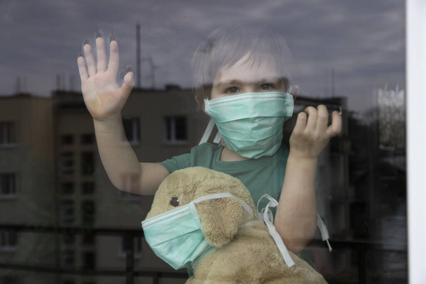 コロナウイルスCovid-19,男の子と彼のぬいぐるみ子犬で保護マスク窓から見て、自宅で隔離  - 写真・画像