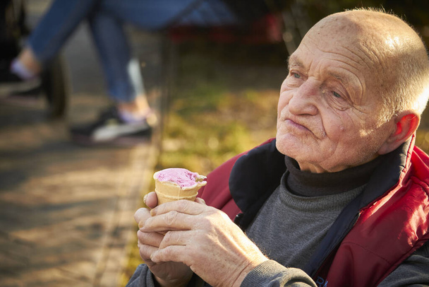 Ογδόντα χρονών άντρας περπατάει σε δημόσιο πάρκο, τρώει παγωτό σε κούπα βάφλα. - Φωτογραφία, εικόνα