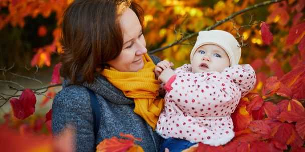 Happy smiling woman with baby girl on sunny fall day in park. Beautiful family of two in bright red foliage. Autumn activities with kids - Foto, afbeelding