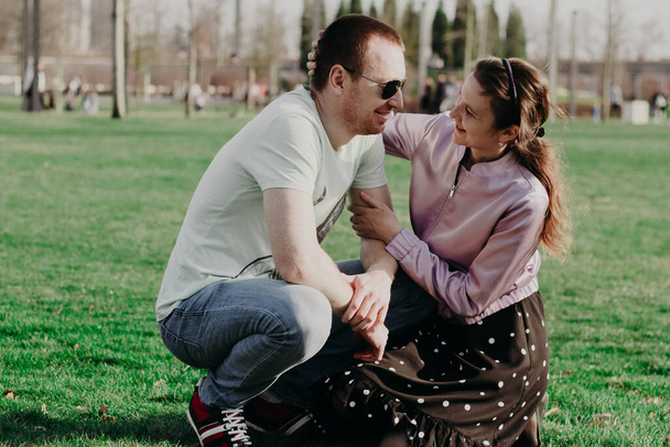 Ευτυχισμένο νεαρό παντρεμένο ζευγάρι στο πάρκο. Ένας άντρας και ένα κορίτσι κάθονται στο γρασίδι ο ένας δίπλα στον άλλο και κοιτάζονται με τρυφερότητα και αγάπη σε μια ηλιόλουστη ανοιξιάτικη μέρα. - Φωτογραφία, εικόνα