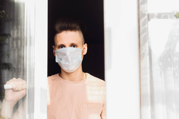 Przystojny młody człowiek w masce ochronnej stoi w domu za oknem. Wezwanie do samodzielnej izolacji z powodu pandemii i ryzyka zakażenia koronawirusem. - Zdjęcie, obraz