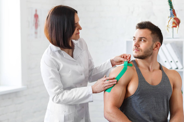 Kinesiology taping. Physical therapist applying kinesiology tape to patient shoulder. Female therapist treating injured shoulder of male athlete. Post traumatic rehabilitation, sport physical therapy - Fotoğraf, Görsel