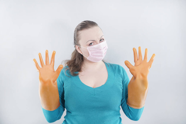 kavkazské ženy středního věku nosí ochrannou lékařskou masku a velké gumové rukavice pro sterilitu na bílém pozadí. žena v domácím svetru je připravena uklidit dům v karanténě - Fotografie, Obrázek