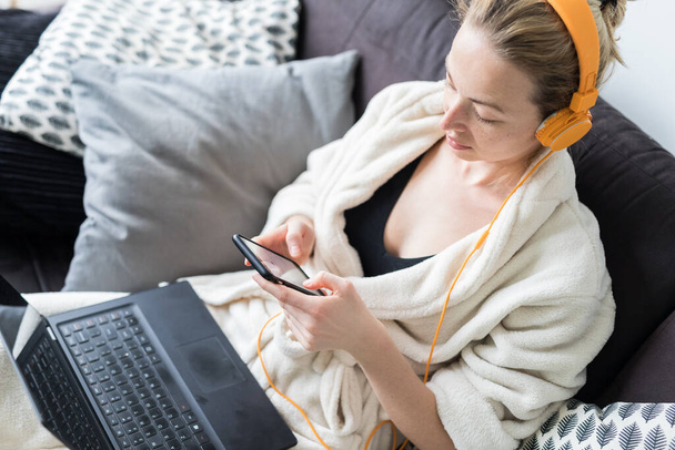 社会的距離。家にいて。バスローブの女性は自宅のソファで快適に、ビデオチャットや彼女の愛する人との接続を維持するために電話でソーシャルメディアアプリを使用しています. - 写真・画像