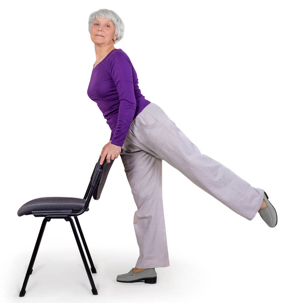 Ευτυχισμένο γοητευτικό όμορφη ηλικιωμένη γυναίκα κάνει ασκήσεις στο σπίτι σε μια καρέκλα. Γυμναστική γυμναστική για την υγεία. Σε λευκό φόντο - Φωτογραφία, εικόνα