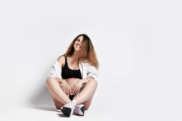 Pregnant mom in black sport shorts and top, shirt, pink sneakers. She is smiling, sitting on floor isolated on white. Copy space - 写真・画像