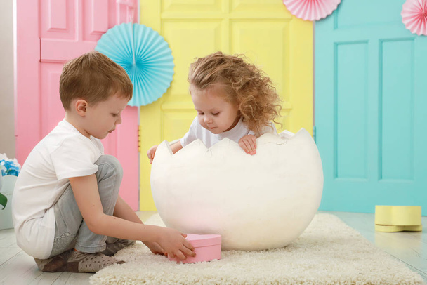Pretty little blonde curly girl is sitting in a white egg and adorable blond boy portrait in white t-shirt on yellow, pink and blue background. Kid gender relations concept. - 写真・画像