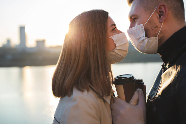 Young couple, a man and a woman in medical masks and gloves, drink coffee from disposable cups on the street and look at each other against the background of the river and the city. Quarantine - Fotoğraf, Görsel