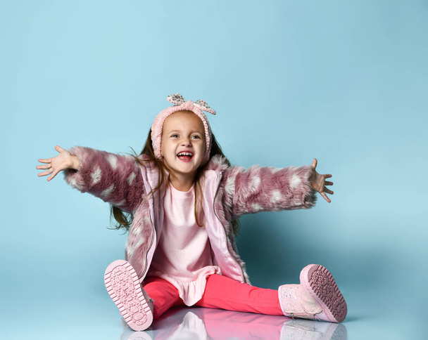 Child in pink faux fur coat, pants and boots. She smiling, gesticulating, sitting on floor against blue background. Close up - Foto, Imagem