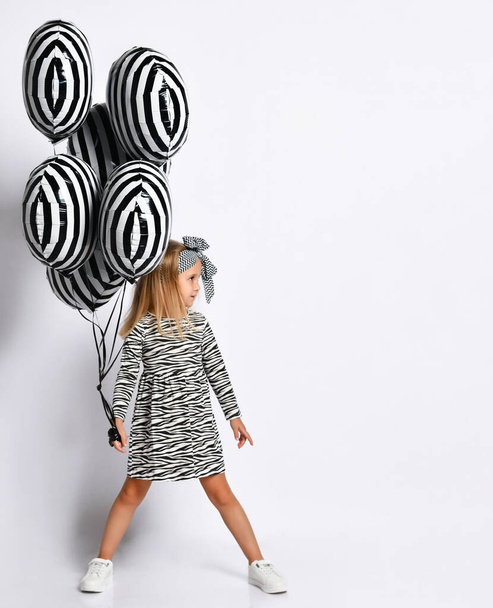Little blonde female in zebra-print dress, headband and sneakers. Holding striped balloons, posing isolated on white. Full length - Фото, изображение