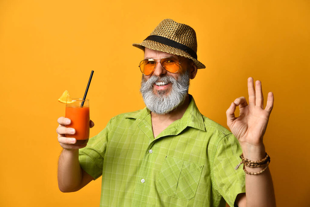 Bearded grandpa in hat, green shirt, sunglasses. Smiling, showing okay sign, holding glass of juice, posing on orange background - Fotó, kép
