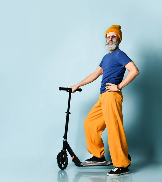 Bearded grandpa in t-shirt, sunglasses, orange pants, hat, gumshoes. Riding black scooter, showing fist, posing on blue background - Photo, image