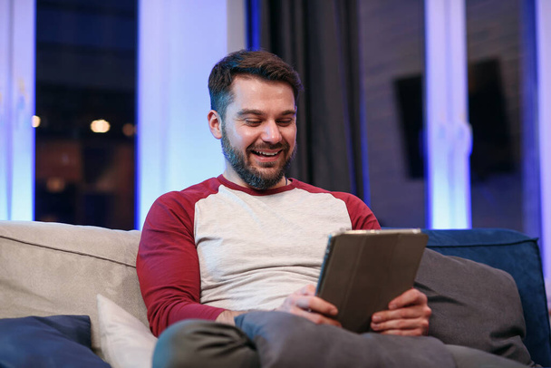 Slow motion of likable modern good-natured 30-aged man with well-groomed beard which resting on the sofa at home and browsing apps on tablet pc with lucky smile - Photo, Image
