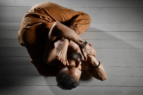 Aged yogi man with naked torso, in brown pants and bracelets is performing his flexibility. Lying on a white wooden floor - Photo, image