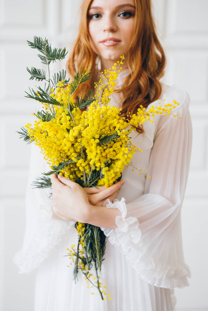 Portrait of a red-haired beautiful girl with a Mimosa in a long white dress. Spring concert. Toning. - Фото, изображение