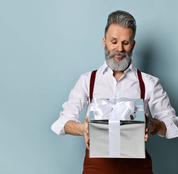 Gray-haired man in white shirt, brown pants and suspenders. Smiling, holding silver gift box, posing against blue background - Photo, Image