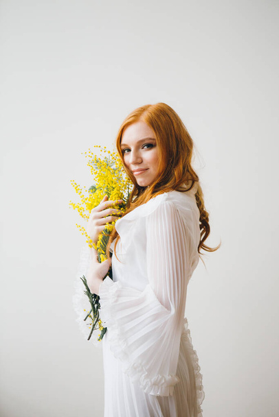 Portrait of a red-haired beautiful girl with a Mimosa in a long white dress. Spring concert. Toning. - Foto, Imagem