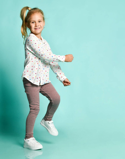 Little blonde kid in shirt with hearts print, checkered pants, white sneakers. Smiling, dancing on blue background. Full length - 写真・画像