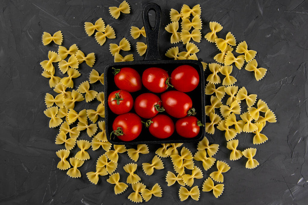 Raw pasta farfalle, tomatoes and frying pan on dark background. Top views, close-up. - Photo, Image