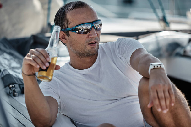 Hırvatistan, Split, 15 Eylül 2019: Arka planda tekneleri olan bir yelkenli yarışının vahşi katılımcısı yaklaşan yarışı bekliyor, bira içiyor, kayakçı veya denizci, güneş gözlüğü - Fotoğraf, Görsel