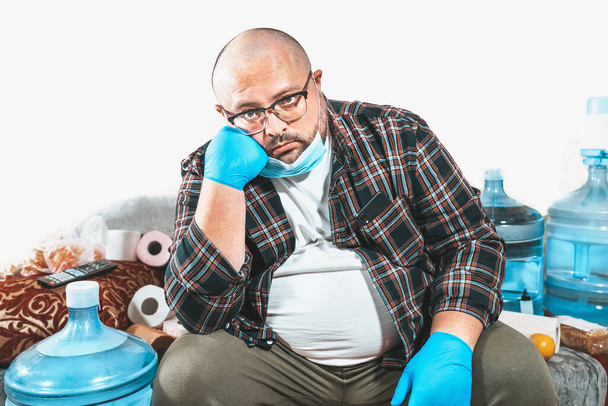 Nudný muž s ochrannou maskou sedí na pohovce s toaletním papírem a láhvemi vody, coronavirus covid-19. Karanténa, sebeizolace a zůstat doma koncept, kopírovat prostor - Fotografie, Obrázek