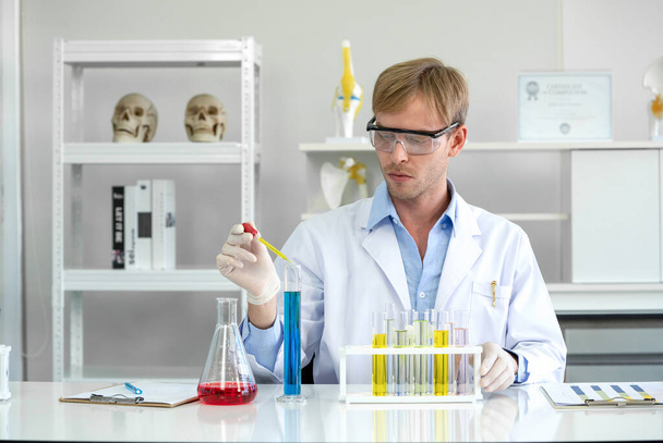 Ein junger brasilianischer Wissenschaftler mit einem Brillenlabor mischt vorsichtig kosmetische Chemikalien zusammen. Arbeitsatmosphäre im Chemielabor. Reagenzgläser mit Chemikalien auf dem Tisch. - Foto, Bild