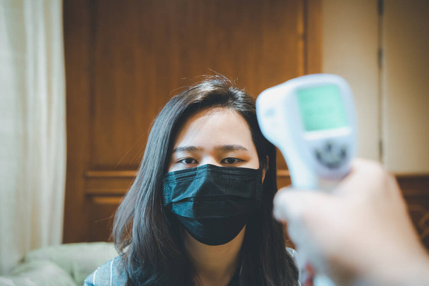 Asiatin mit Maske Atemschutzmaske gegen epidemische Grippe Covid19 oder Coronavirus und Körpertemperatur Gesundheitscheck messen Fieberwerte mit digitalem Infrarot-Thermometer für das Gesundheitswesen - Foto, Bild