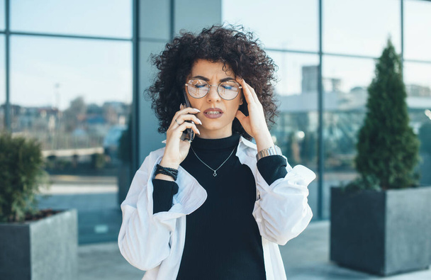 Figyelmes fehér hölgy göndör hajjal és szemüveggel, megérintve a haját és pózolva egy üzleti telefonbeszélgetés közben. - Fotó, kép