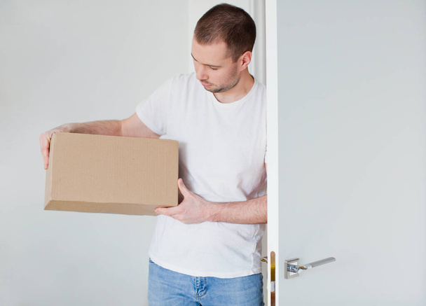 Caucasian man with cardboard box opens the door and enters the room. Delivery man on a white background - Foto, Imagem