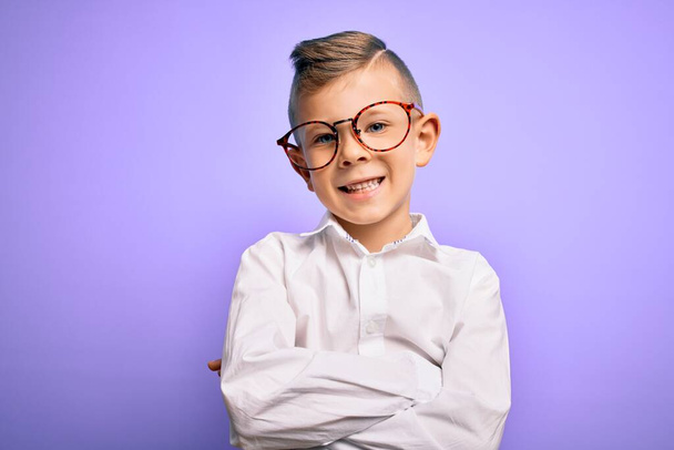 Ein kleines kaukasisches Kind mit blauen Augen, Brille und weißem Hemd vor violettem Hintergrund, das mit verschränkten Armen in die Kamera lächelt. Positiver Mensch. - Foto, Bild