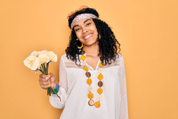 Genç Afro-Amerikan kıvırcık saçlı, güneş gözlüğü takan, elinde bir buket çiçek tutan, mutlu yüzlü, kendine güvenen, gülümseyen, dişlerini gösteren bir kadın. - Fotoğraf, Görsel