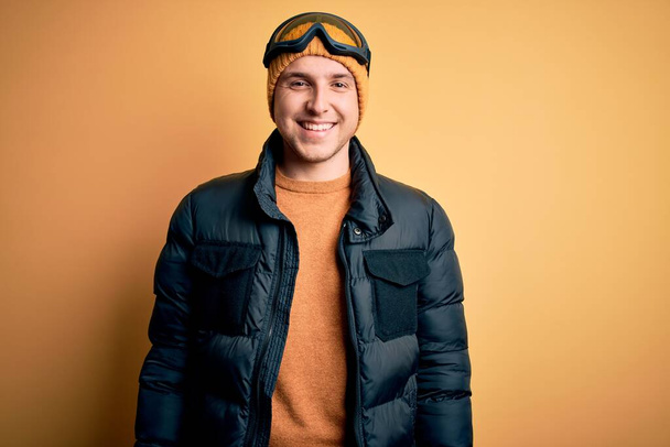 Νεαρός όμορφος καυκάσιος άνδρας με καπέλο, παλτό και γυαλιά σκι για χειμώνα και χιόνι με χαρούμενο και δροσερό χαμόγελο στο πρόσωπο. Τυχερός άνθρωπος. - Φωτογραφία, εικόνα