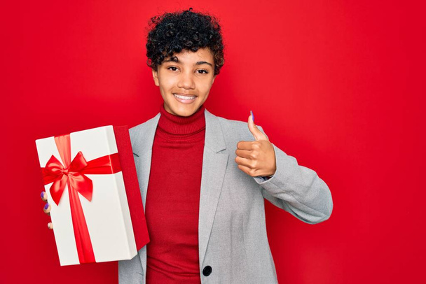 Beautiful african american afro business woman holding gift over isolated red background happy with big smile doing ok sign, thumb up with fingers, excellent sign - Fotó, kép