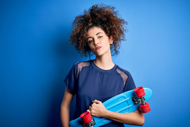 Young sporty woman with curly hair and piercing holding skate over blue background with a confident expression on smart face thinking serious - Photo, image