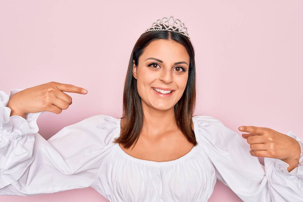 Young beautiful brunette woman wearing princess tiara standing over isolated pink background looking confident with smile on face, pointing oneself with fingers proud and happy. - Photo, Image