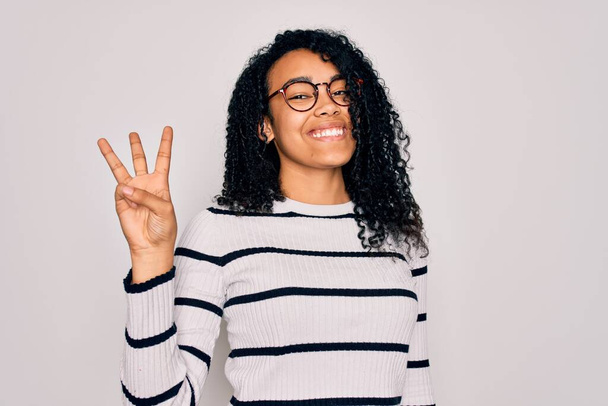 Молодая африканская американка в полосатом свитере и очках на белом фоне показывает и указывает пальцами номер три, улыбаясь уверенно и счастливо
. - Фото, изображение