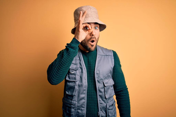 Handsome tourist man with beard on vacation wearing explorer hat over yellow background doing ok gesture shocked with surprised face, eye looking through fingers. Unbelieving expression. - Photo, Image