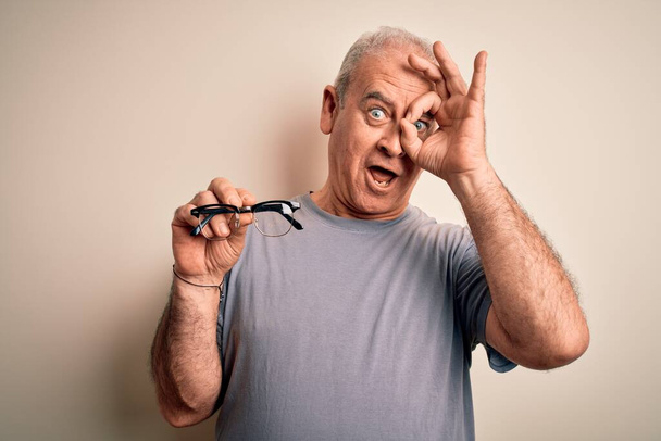 Středního věku chudokrevný optický muž drží brýle na izolovaném bílém pozadí se šťastnou tváří usmívá dělá v pořádku znamení s rukou na oku dívá skrz prsty - Fotografie, Obrázek