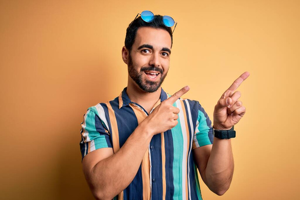 Όμορφος τουρίστας με γενειάδα σε διακοπές φορώντας καλοκαιρινό ριγέ πουκάμισο και γυαλιά ηλίου χαμογελώντας και κοιτάζοντας την κάμερα με τα δύο χέρια και τα δάχτυλα στο πλάι. - Φωτογραφία, εικόνα
