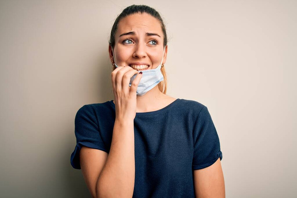Beautiful blonde woman with blue eyes wearing medical mask over white background looking stressed and nervous with hands on mouth biting nails. Anxiety problem. - Foto, Bild