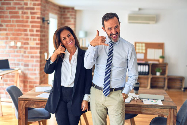 Двое бизнесменов среднего возраста, стоящих вместе на встрече в офисе и улыбающихся, делают телефонный жест руками и пальцами, как будто разговаривают по телефону. Коммуникационные концепции
. - Фото, изображение