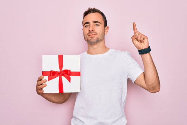 Mladý pohledný muž s modrýma očima drží dárek k narozeninám nad izolovaným růžovým pozadím překvapený nápadem nebo otázkou ukazující prstem se šťastnou tváří, číslo jedna - Fotografie, Obrázek