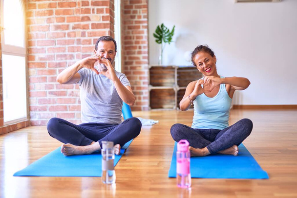 Sportliches Paar mittleren Alters, das auf einer Matte sitzt und Stretching-Yoga-Übungen im Fitnessstudio macht und verliebt lächelt und Herzsymbol und Form mit den Händen zeigt. Romantisches Konzept. - Foto, Bild