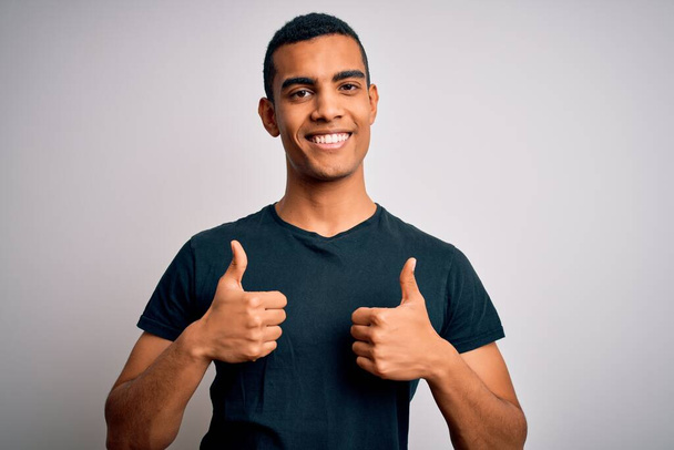 Ein junger gutaussehender afrikanisch-amerikanischer Mann in lässigem T-Shirt steht über einem weißen Erfolgsschild und macht eine positive Geste mit der Hand, Daumen hoch lächelnd und glücklich. Fröhliche Miene und Siegergeste. - Foto, Bild