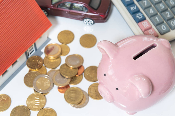 Pinkfarbenes Sparschwein und ein Haufen Geldmünzen, Autos, Häuser und Taschenrechner. Finanzkonzepte und Sparen. - Foto, Bild