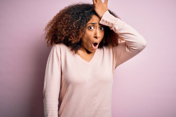 Jonge Afro-Amerikaanse vrouw met afro haar draagt casual trui over roze achtergrond verrast met de hand op het hoofd voor fout, herinner fout. Vergeten, slecht geheugen concept. - Foto, afbeelding