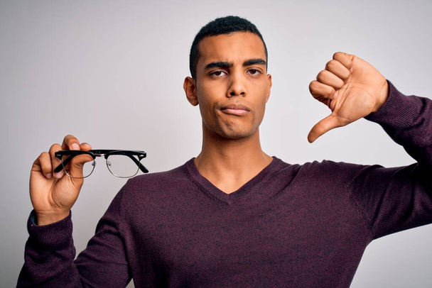 Jovem homem óptico afro-americano bonito segurando óculos sobre fundo branco isolado com rosto zangado, sinal negativo mostrando antipatia com os polegares para baixo, conceito de rejeição
 - Foto, Imagem