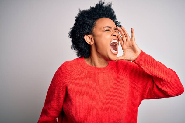 Jonge mooie Afro-Amerikaanse afro vrouw met krullend haar draagt rode casual trui schreeuwen en schreeuwen luid naar zij met de hand op de mond. Communicatieconcept. - Foto, afbeelding