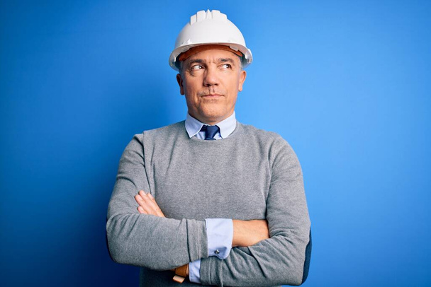 Moyen âge beau ingénieur aux cheveux gris homme portant un casque de sécurité sur fond bleu souriant regardant vers le côté et regardant loin en pensant
. - Photo, image